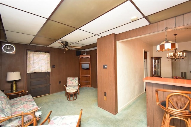 living room featuring ceiling fan with notable chandelier, a paneled ceiling, indoor bar, wood walls, and carpet flooring