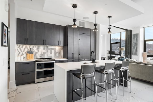 kitchen featuring decorative backsplash, a kitchen island with sink, range with two ovens, pendant lighting, and sink