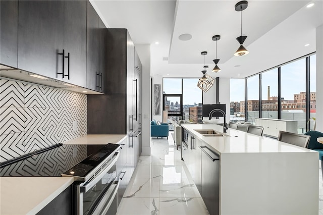 kitchen featuring stainless steel range with electric stovetop, a center island with sink, a wall of windows, pendant lighting, and sink