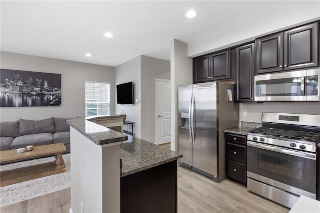 kitchen with dark brown cabinets, appliances with stainless steel finishes, dark stone countertops, and light hardwood / wood-style floors