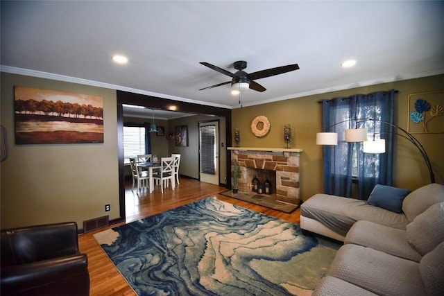 living room with ceiling fan, crown molding, wood-type flooring, and a stone fireplace