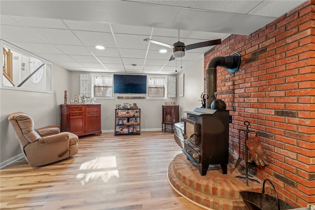 living room with light wood-type flooring, ceiling fan, a drop ceiling, and a wood stove