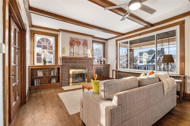 living room featuring wood-type flooring, beamed ceiling, a fireplace, ceiling fan, and crown molding