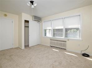 unfurnished bedroom with radiator heating unit, a wall mounted air conditioner, a closet, and light colored carpet