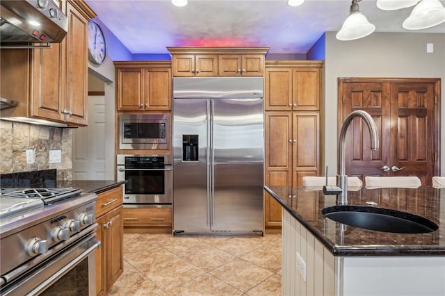 kitchen with decorative light fixtures, dark stone counters, built in appliances, sink, and range hood