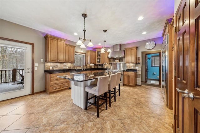 kitchen with wall chimney exhaust hood, a kitchen breakfast bar, hanging light fixtures, backsplash, and a kitchen island with sink