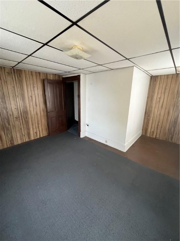 basement with a drop ceiling, wood walls, and dark colored carpet