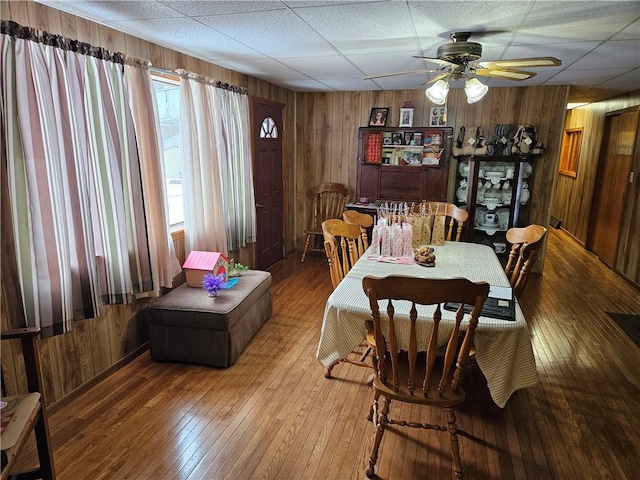 dining area with ceiling fan, wooden walls, wood-type flooring, and a paneled ceiling