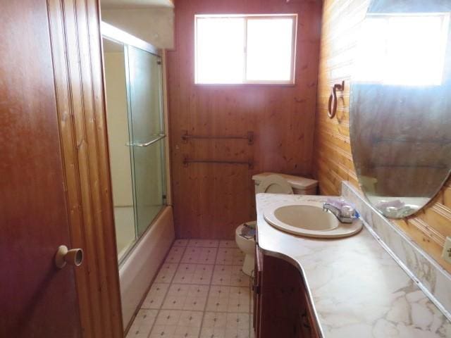 full bathroom featuring vanity, bath / shower combo with glass door, toilet, and wood walls