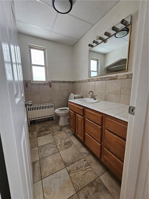 bathroom with toilet, tile walls, radiator heating unit, vanity, and a drop ceiling