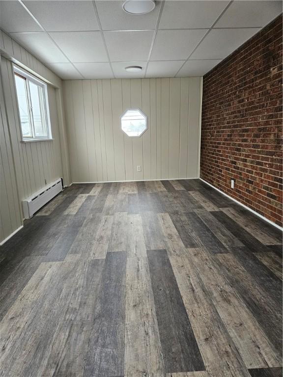 empty room featuring a baseboard radiator, brick wall, dark hardwood / wood-style flooring, and a drop ceiling