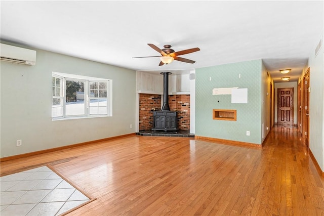 unfurnished living room with ceiling fan, a wood stove, a wall unit AC, and light hardwood / wood-style floors