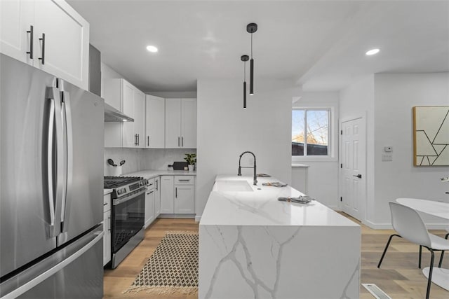 kitchen featuring pendant lighting, wall chimney range hood, appliances with stainless steel finishes, white cabinetry, and a center island with sink