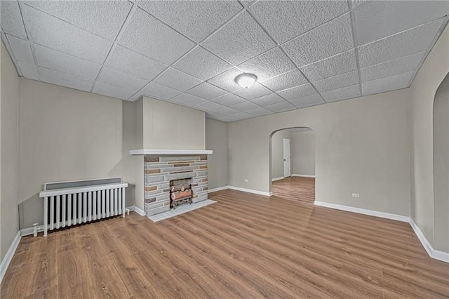 unfurnished living room with arched walkways, a stone fireplace, radiator, and wood finished floors