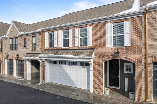 townhome / multi-family property with a garage, brick siding, driveway, and roof with shingles