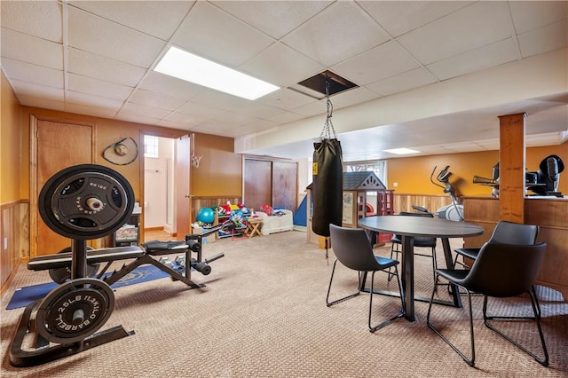 exercise room with a paneled ceiling, light colored carpet, and wood walls