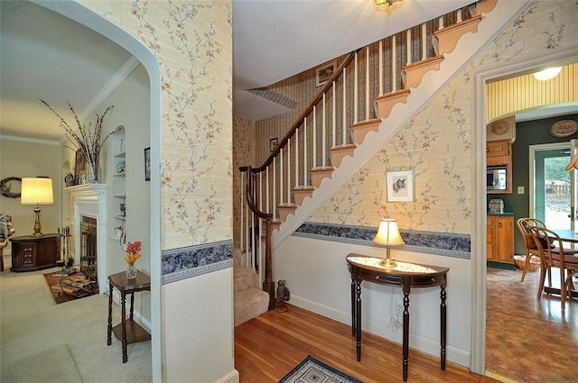 staircase with wallpapered walls, crown molding, wood finished floors, and arched walkways