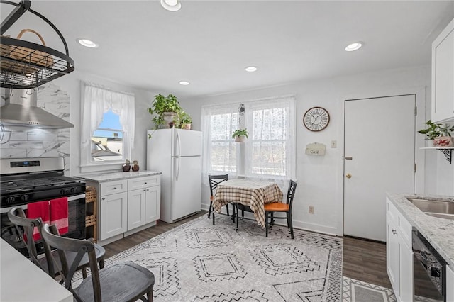 kitchen featuring wall chimney range hood, stainless steel gas range oven, light stone countertops, white cabinets, and white fridge