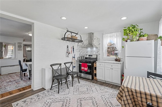 kitchen with stainless steel gas stove, white cabinetry, hardwood / wood-style flooring, white fridge, and wall chimney range hood