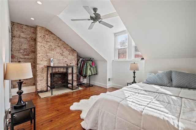 bedroom with vaulted ceiling, a brick fireplace, hardwood / wood-style flooring, ceiling fan, and brick wall