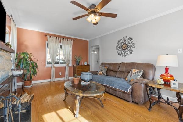 living room featuring ceiling fan, ornamental molding, wood-type flooring, and a stone fireplace