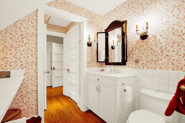 bathroom with wood finished floors, vanity, a wainscoted wall, toilet, and wallpapered walls
