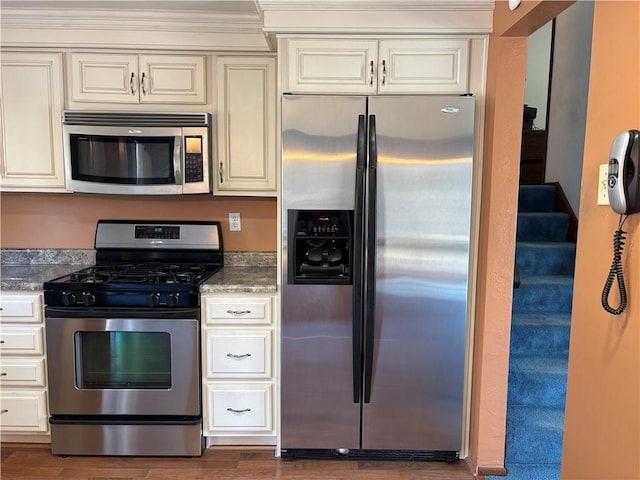 kitchen with stainless steel appliances, cream cabinetry, dark wood finished floors, and dark stone countertops