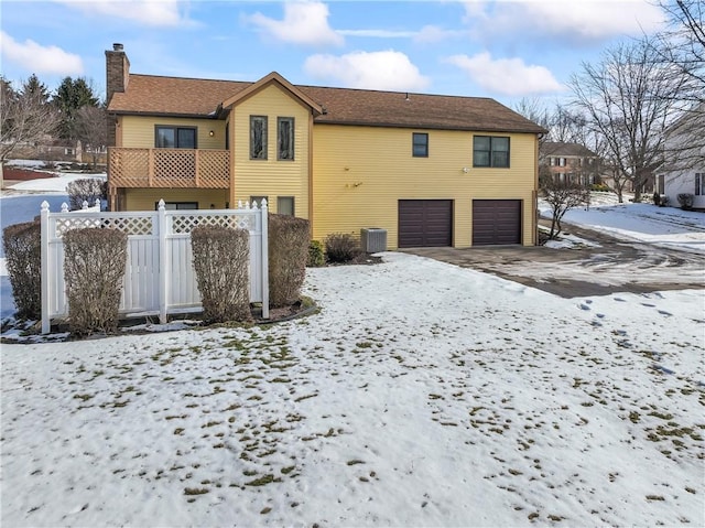 snow covered back of property with central AC, a balcony, and a garage