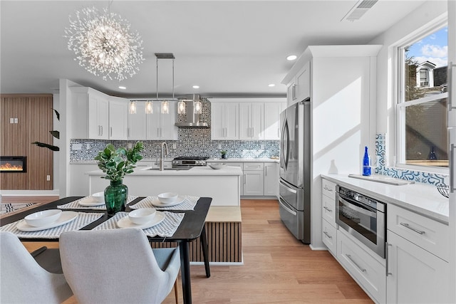 kitchen with decorative light fixtures, white cabinets, light hardwood / wood-style floors, stainless steel appliances, and wall chimney range hood