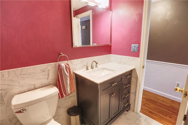 half bath with wainscoting, toilet, marble finish floor, vanity, and tile walls