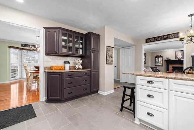 kitchen featuring a brick fireplace, dark brown cabinets, a notable chandelier, decorative backsplash, and white cabinets