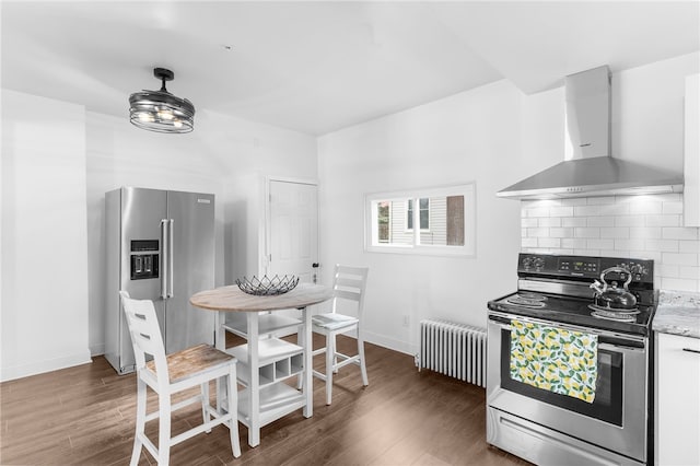 kitchen with wall chimney exhaust hood, dark hardwood / wood-style flooring, radiator, stainless steel appliances, and decorative backsplash