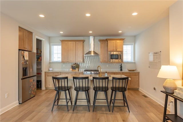 kitchen featuring wall chimney range hood, stainless steel appliances, tasteful backsplash, an island with sink, and light wood-type flooring