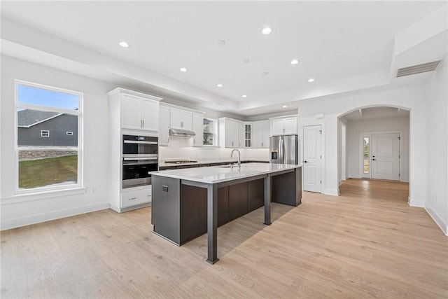 kitchen featuring arched walkways, appliances with stainless steel finishes, a center island with sink, and white cabinets