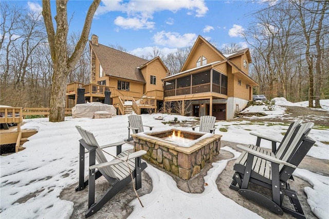 snow covered house featuring a deck, a sunroom, a fire pit, and an attached garage