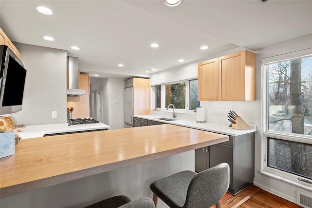 kitchen featuring butcher block countertops, wall chimney range hood, sink, kitchen peninsula, and light brown cabinets