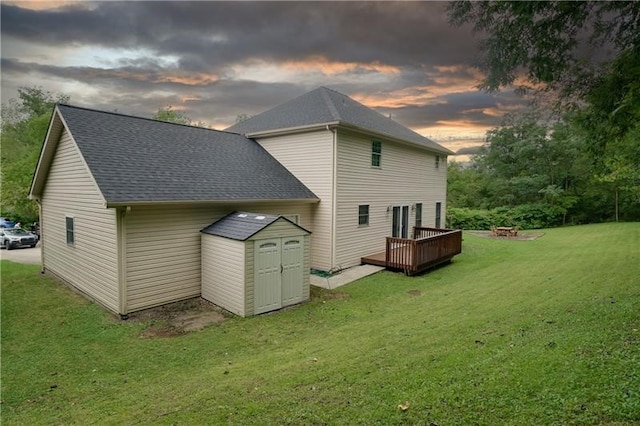 back house at dusk featuring a wooden deck, a fire pit, a shed, and a lawn