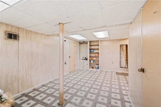 basement with built in shelves, a drop ceiling, and wood walls