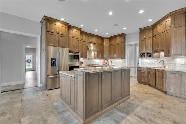 kitchen featuring wall chimney range hood, sink, a kitchen island with sink, stainless steel appliances, and light stone countertops