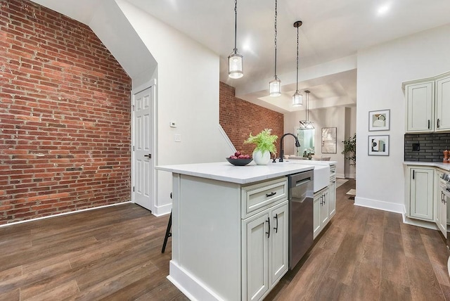 kitchen with dark wood-type flooring, hanging light fixtures, a center island with sink, dishwasher, and brick wall