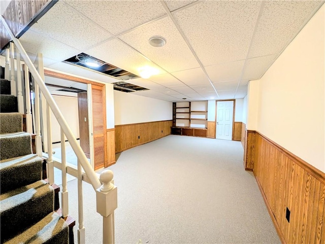 basement featuring a drop ceiling, light colored carpet, and wood walls