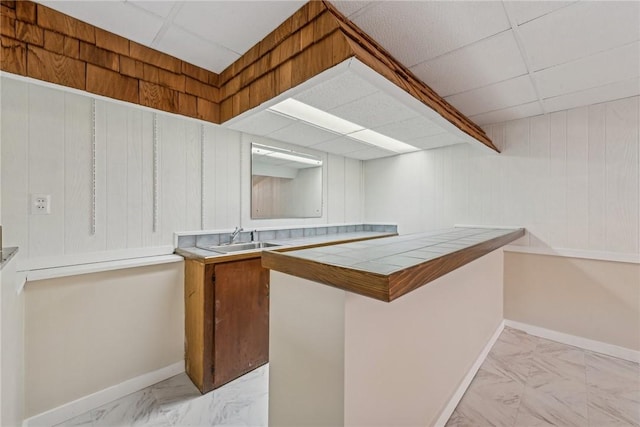 bar with sink, tile countertops, wood walls, and a paneled ceiling