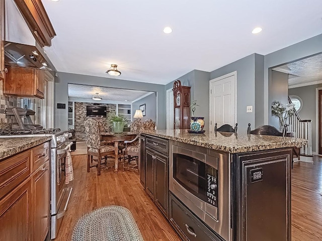 kitchen with appliances with stainless steel finishes, range hood, light stone counters, light hardwood / wood-style floors, and a center island with sink