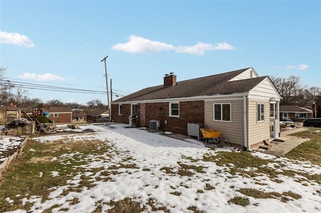 snow covered back of property featuring central AC unit, brick siding, and a chimney