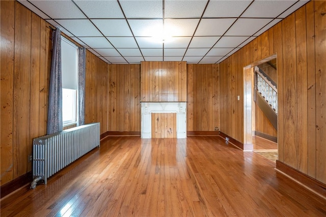 unfurnished living room with a paneled ceiling, radiator heating unit, wooden walls, and hardwood / wood-style floors