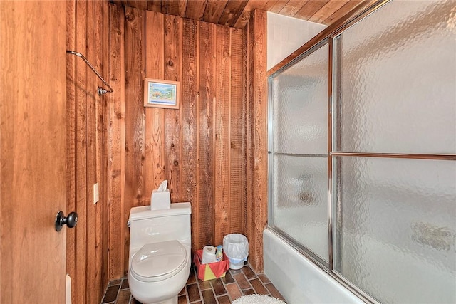 bathroom featuring toilet, combined bath / shower with glass door, and wood walls