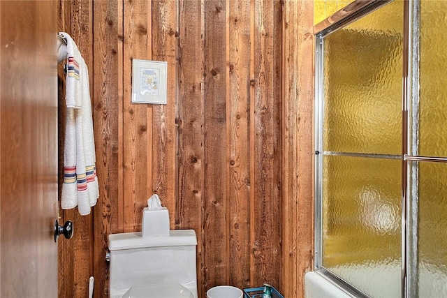 bathroom featuring bath / shower combo with glass door, wooden walls, and toilet