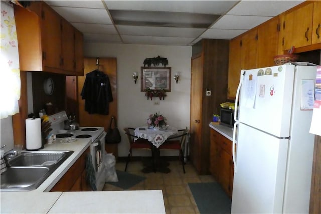 kitchen with a paneled ceiling, light countertops, brown cabinetry, a sink, and white appliances