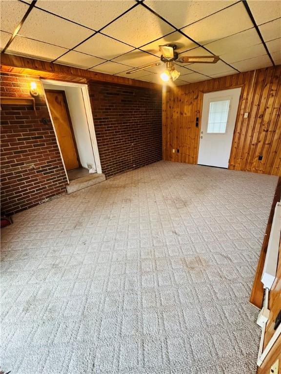 unfurnished living room featuring brick wall, a drop ceiling, ceiling fan, and wood walls