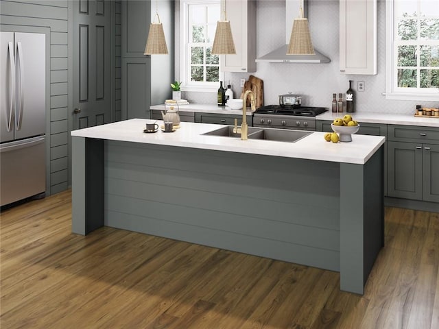 kitchen with gray cabinetry, wall chimney exhaust hood, decorative light fixtures, stainless steel fridge, and a kitchen island with sink